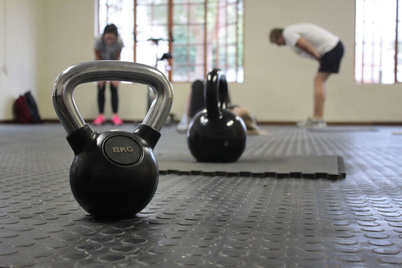 Kettlebells de 8 kg em primeiro plano e alunos treinando ao fundo em sala de treinamento funcional.